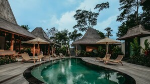 A pool area with lounge chairs and umbrellas, surrounded by wooden deck and tropical plants.