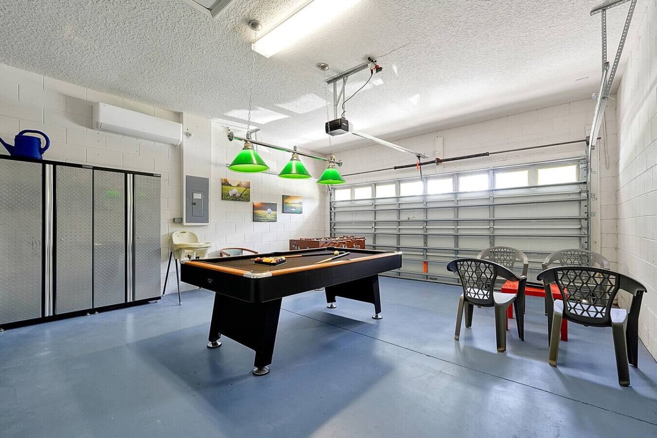 Een pooltafel met groene lampen, een tafelvoetbaltafel en stoelen in een ruimte met een garage deur.