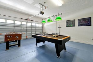 A billiards table with a pool cue, a foosball table, and green pendant lights in a room with a blue floor and white walls.