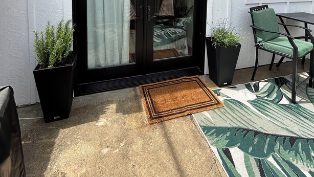 Uma casa com porta de correr de vidro com moldura preta, duas lanternas pretas fixadas na parede, uma cadeira verde e uma pequena mesa.