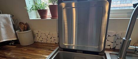 A stainless steel dishwasher in a kitchen with a window view, wooden flooring, and a stone wall.