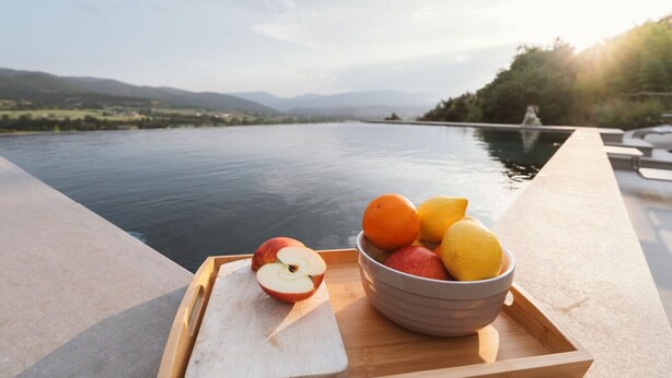 Una bandeja de madera con un bol de fruta y una manzana cortada por la mitad sobre un borde de hormigón junto al agua.
