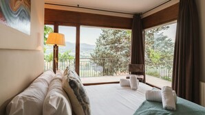Un dormitorio con una cama grande, una silla, un balcón con vistas a árboles y montañas, y un cuadro en la pared.
