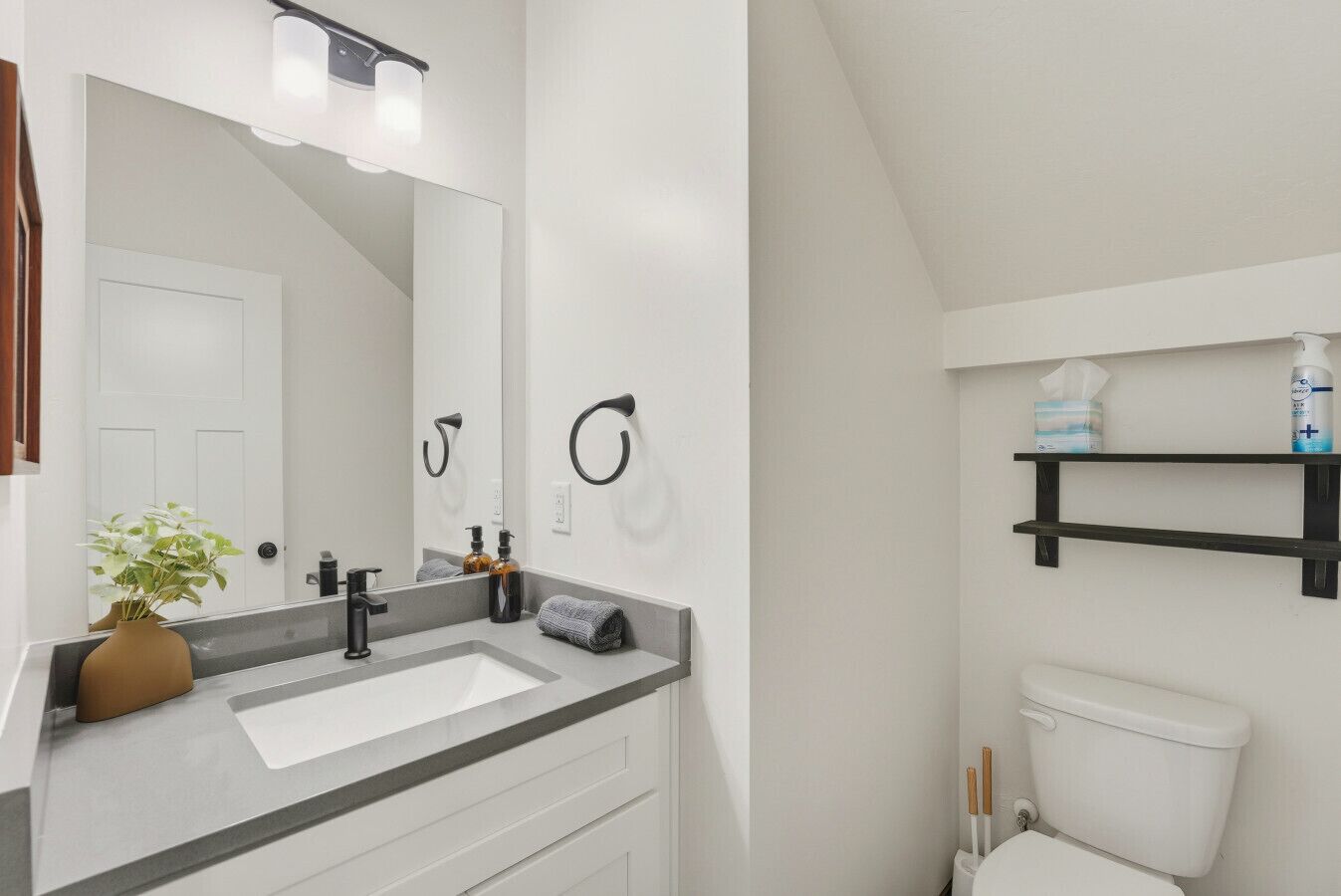 A modern bathroom with a white sink, black faucet, and a large mirror. There is a towel rack, a shelf with toiletries, and a toilet.
