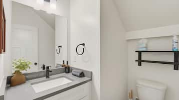A modern bathroom with a white sink, black faucet, and a large mirror. There is a towel rack, a shelf with toiletries, and a toilet.