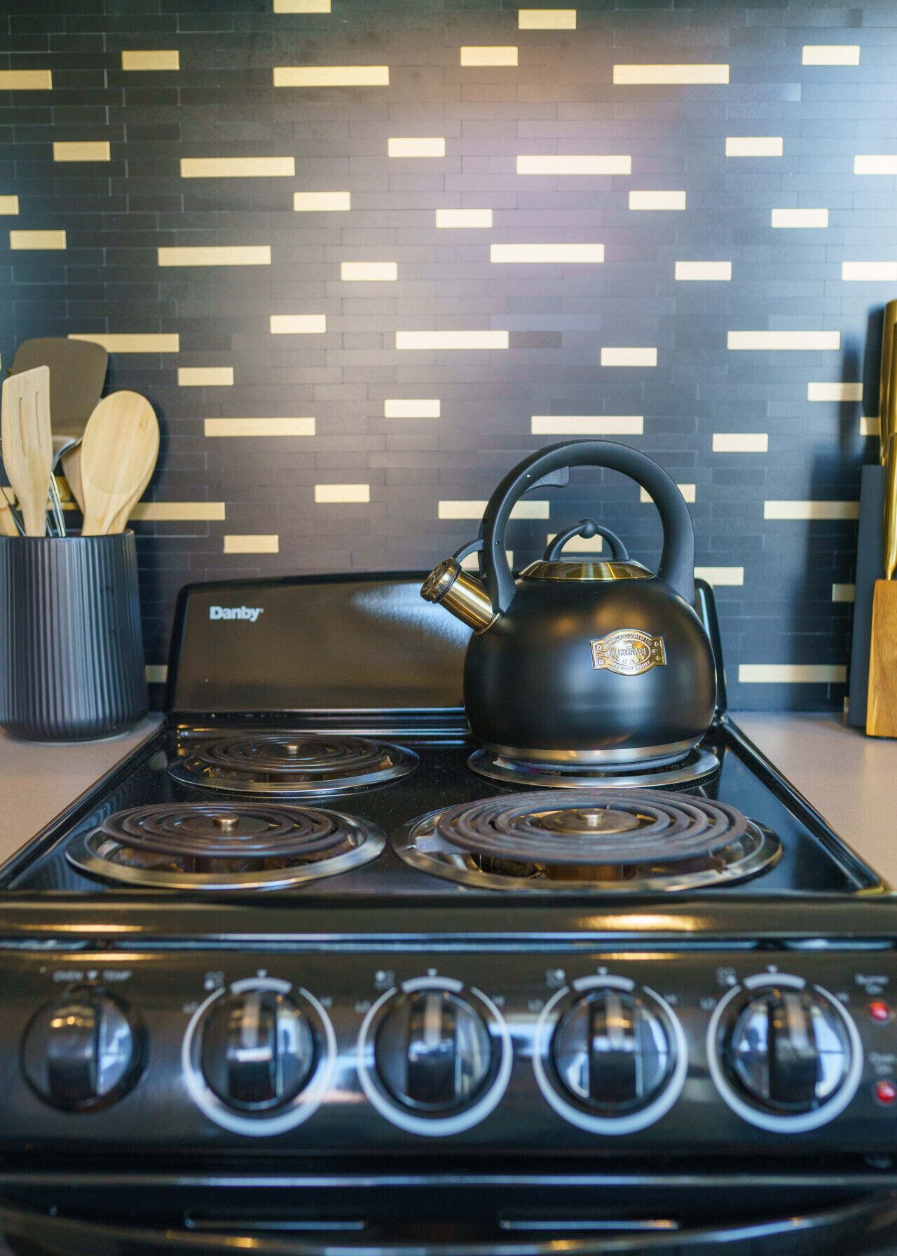 A modern kitchen with a black stove, a kettle, and wooden utensils in a black container.
