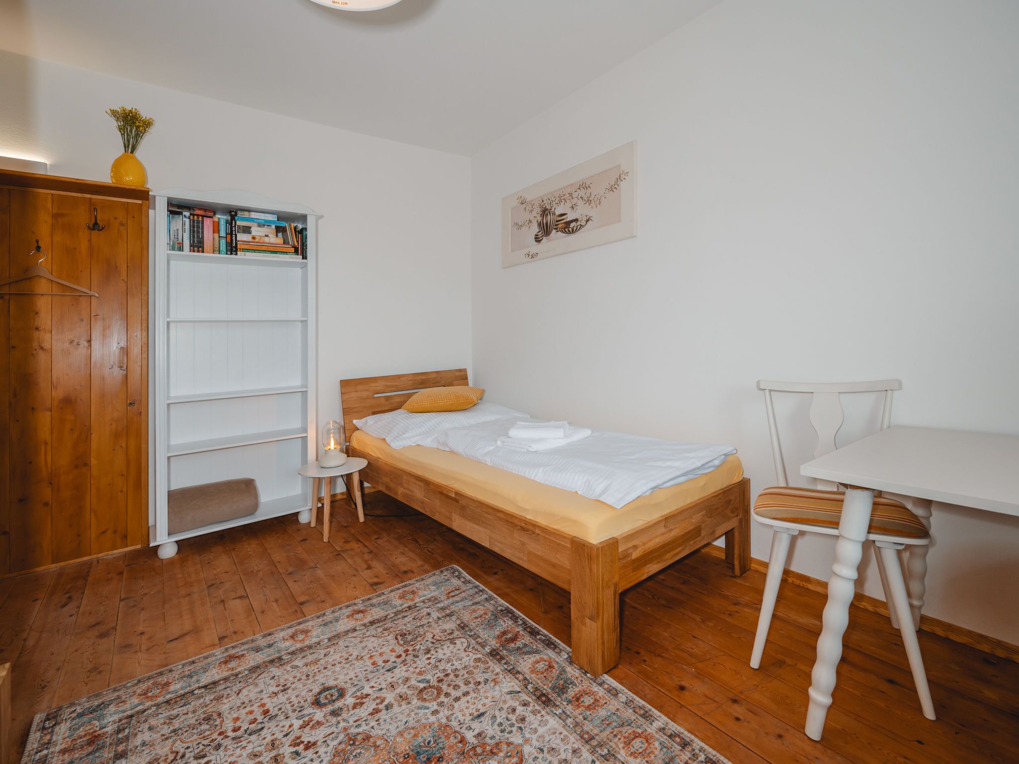 A small, neatly organized bedroom with a wooden bed, a white desk, a chair, a bookshelf, and a rug.