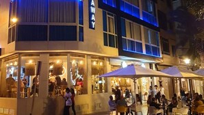 Een hotel met een moderne gevel, verlicht met blauwe neonverlichting, en buiten zitgedeeltes voor gasten.