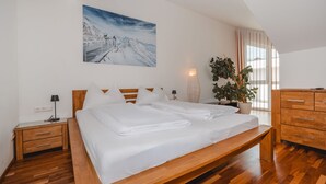Une chambre à coucher moderne avec un lit en bois, une table de chevet, une lampe de chevet, une grande fenêtre avec des rideaux et un tableau accroché au mur représentant un paysage de montagne enneigé.