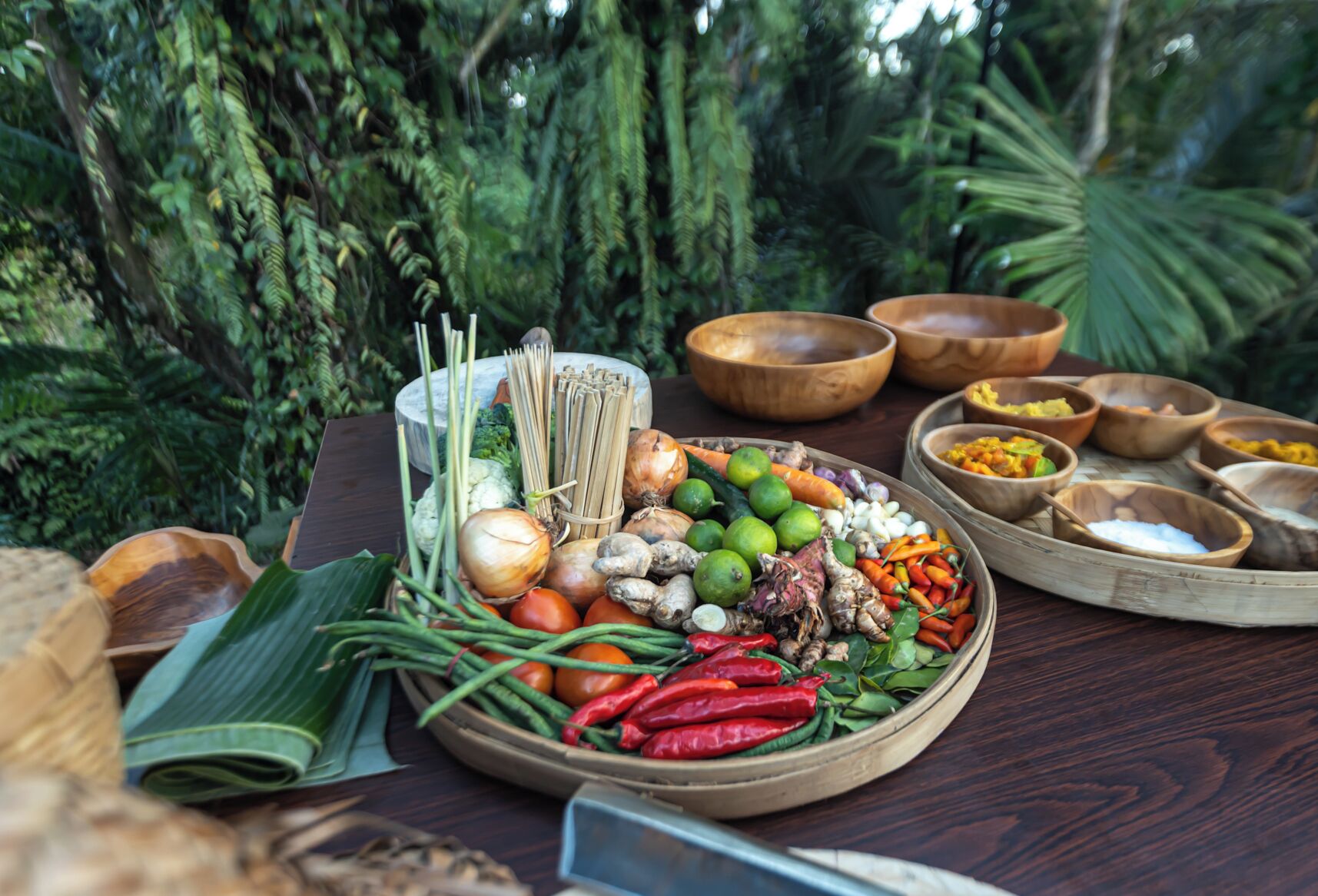 Una selezione di verdure fresche e spezie disposte su un vassoio di legno, con uno sfondo naturale di vegetazione.
