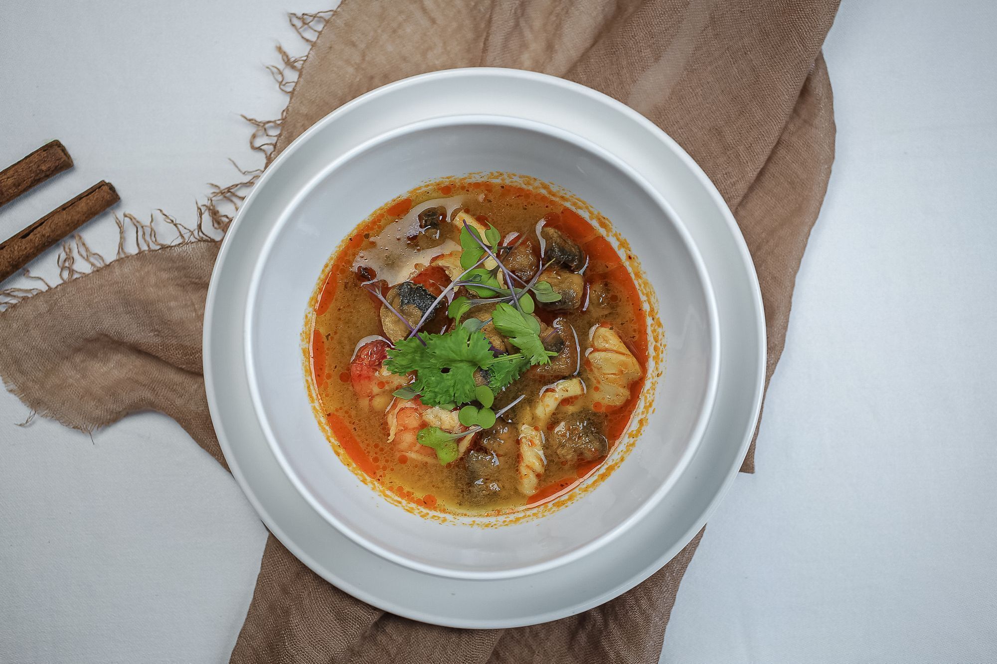 A bowl of soup with various ingredients, placed on a white plate with a brown napkin underneath.