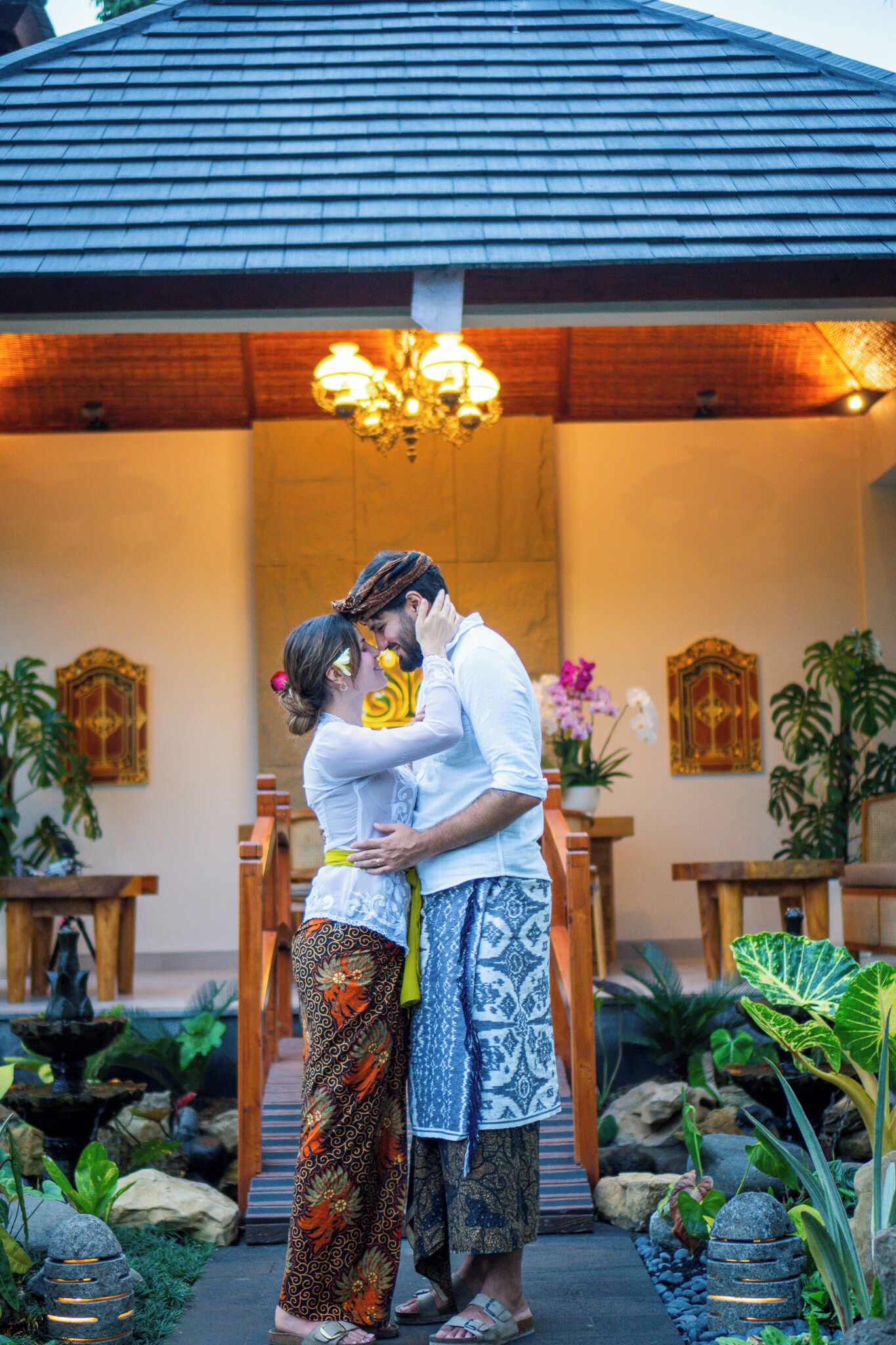 A couple embracing in a traditional setting with a wooden structure and a fountain in the background.