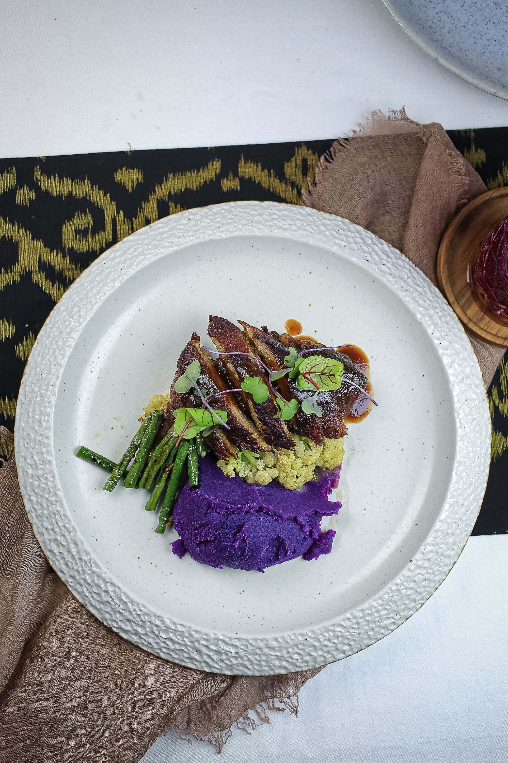 Een wit bord met een gelaagd gerecht van paarse puree, groene bonen en vlees, met een klein schaaltje saus erbij.