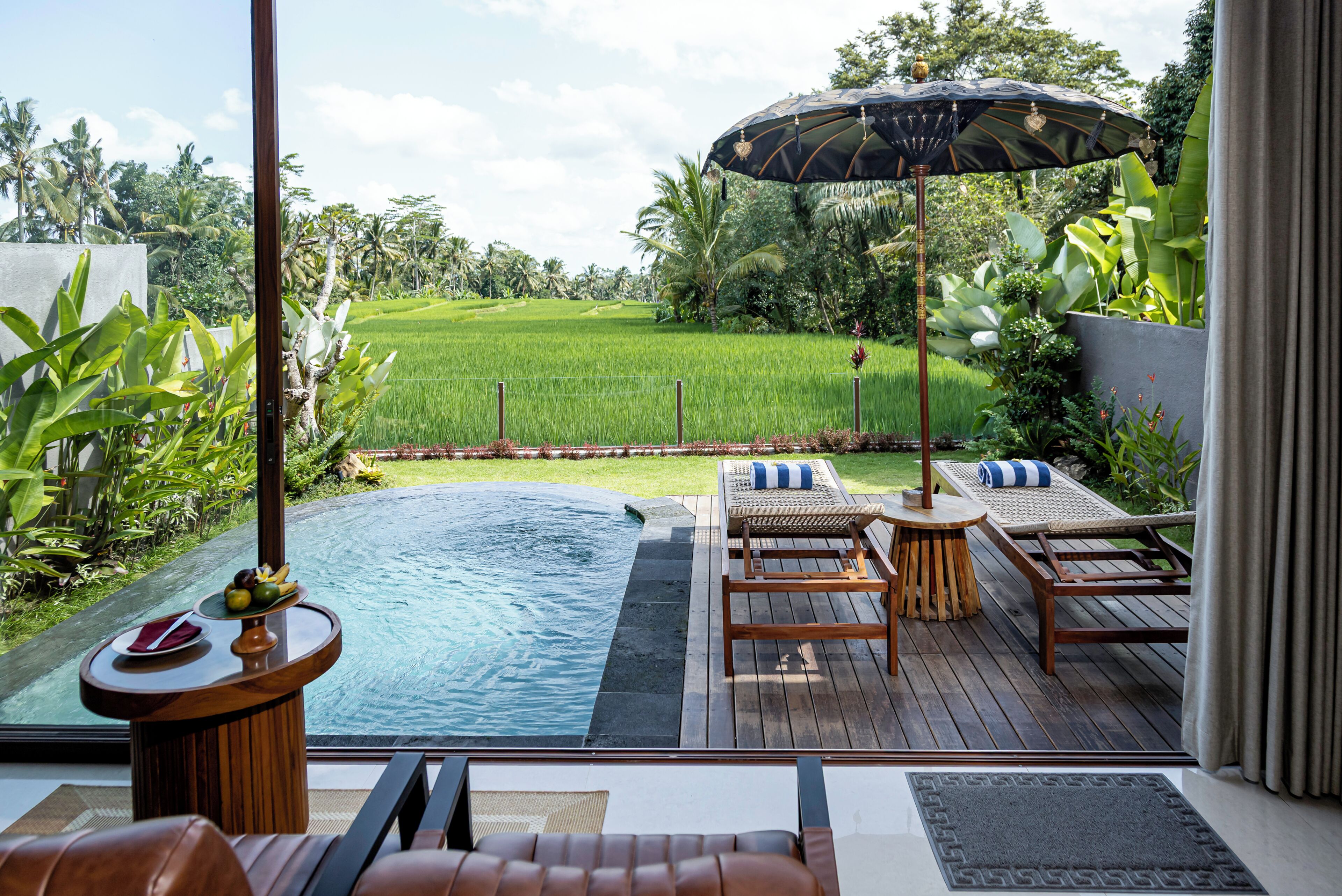 Zona de piscina con vistas a un campo verde exuberante, sillones de madera para tomar el sol y una pequeña mesa con fruta.