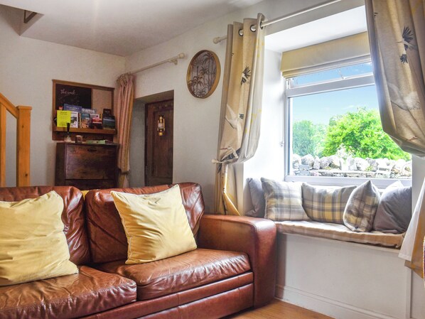 Una sala de estar acogedora con un sofá de cuero marrón, una ventana que da a zonas verdes y una estantería con diversos objetos.