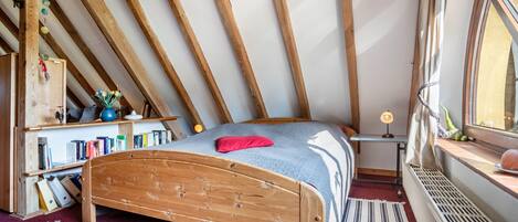 Ein Zimmer mit schrägem Dach, Holzbalken, einem Bett mit blauer Decke, einem kleinen Tisch mit Lampe und einem Fenster mit Blick.