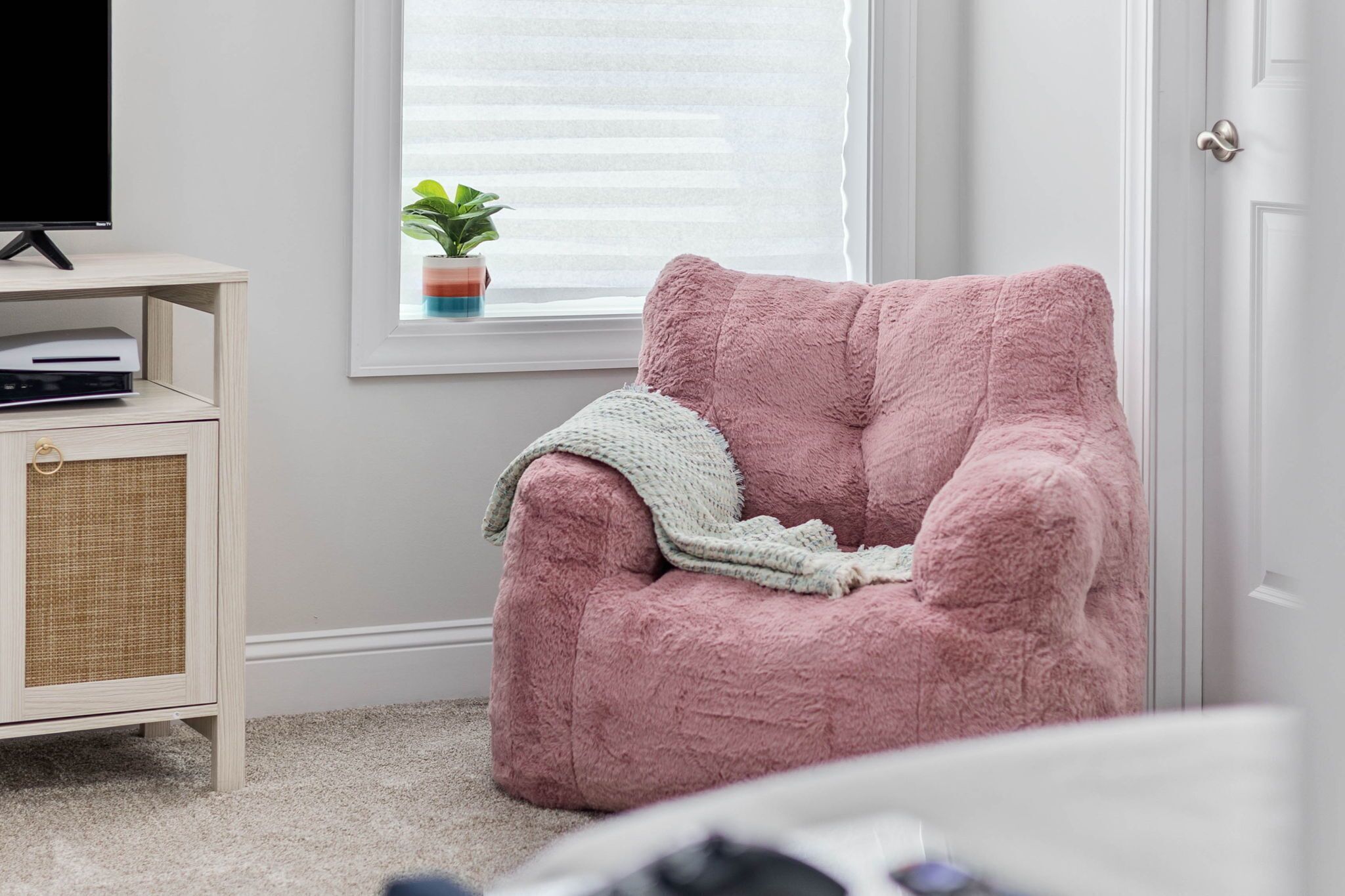 Una habitació acollidora amb una butaca rosa i mullada, un petit armari amb frontal trenat, un televisor sobre un suport i una finestra amb persianes i una planta.