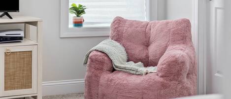 Una habitació acollidora amb una butaca rosa i mullada, un petit armari amb frontal trenat, un televisor sobre un suport i una finestra amb persianes i una planta.