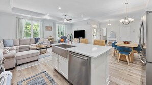 Uma cozinha moderna com uma ilha central, uma sala de jantar com mesa redonda e cadeiras, uma sala de estar com sofá e mesa de centro, e uma grande janela com cortinas.