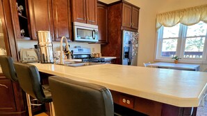 Una cocina con una isla central, taburetes de bar, armarios de madera, microondas, refrigerador y una ventana con cortinas.