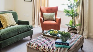 Un salon avec un canapé vert, un fauteuil orange, un pouf à motifs, une plante en pot et un plateau contenant des fruits et des livres.