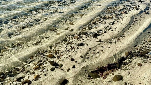 Ein Strand mit klarem Wasser, sichtbaren Sandstrukturen und vereinzelten Felsen.
