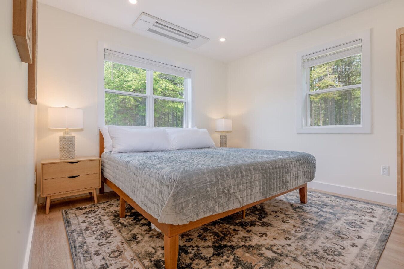 Un dormitorio con una cama, dos mesitas de noche, dos lámparas, un ventanal grande y una alfombra estampada.