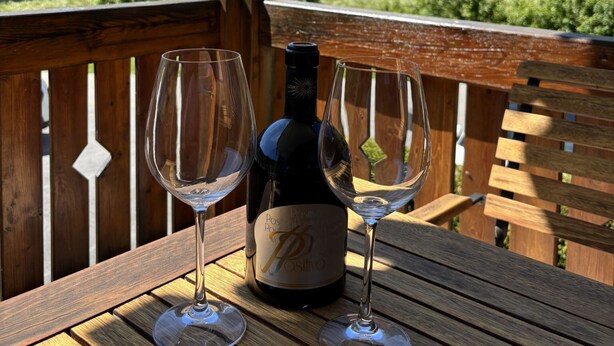 Une table en bois, une bouteille de vin et deux verres à vin vides sont posés sur un balcon qui offre une vue sur un paysage montagneux.