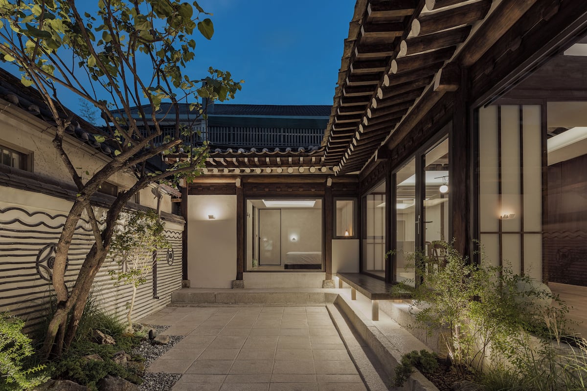 Un edificio tradicional japonés con fachada de madera, un acceso con tejas y un pequeño jardín con un árbol y plantas.