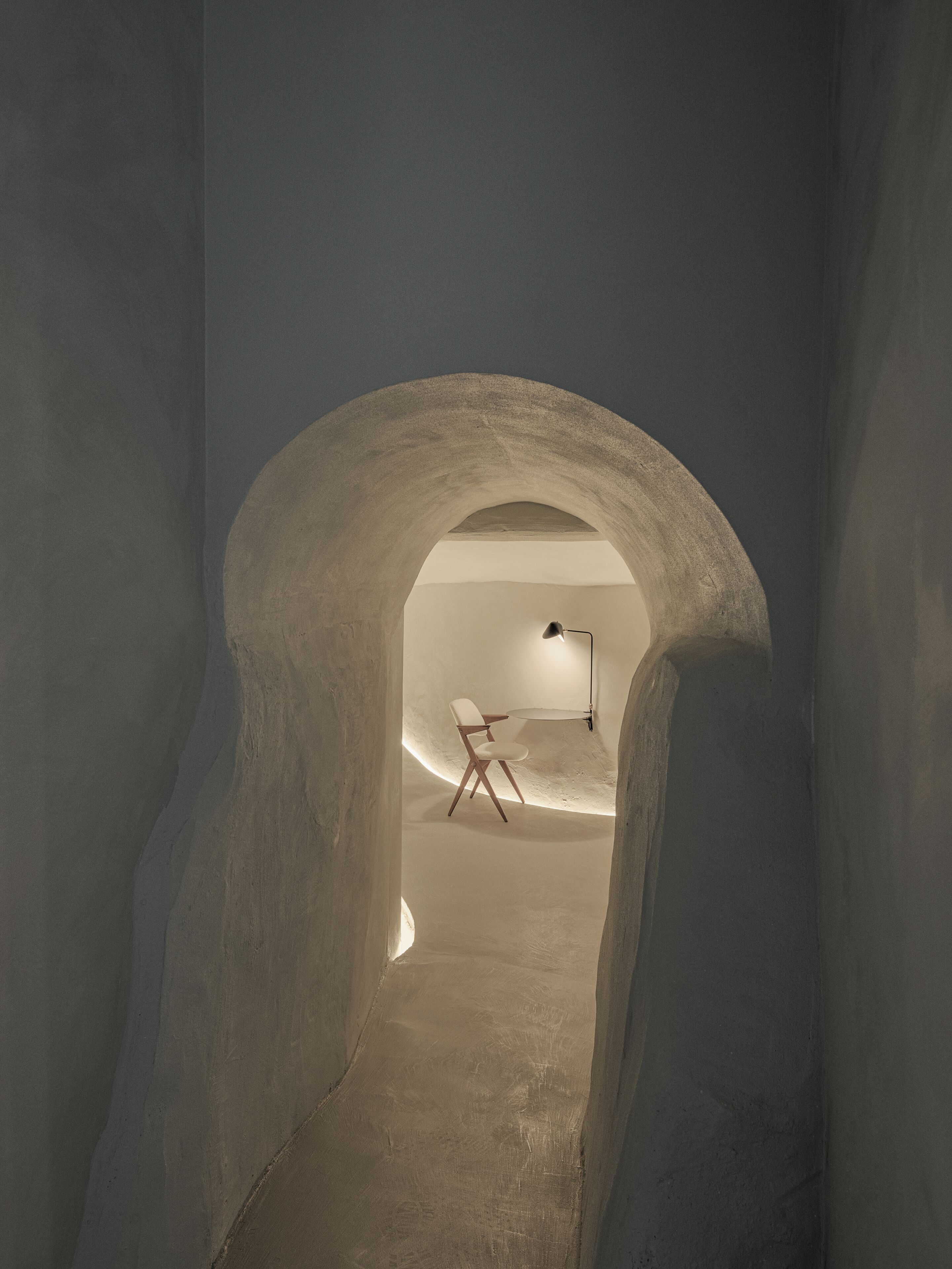 A narrow passageway with a high, arched ceiling leading to a room with a white chair and a desk.