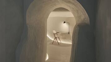 A narrow passageway with a high, arched ceiling leading to a room with a white chair and a desk.