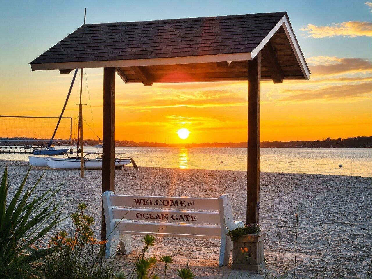 Een houten prieel met een bord waarop 'Welcome to Ocean Gate' staat, bij zonsondergang, met boten op de achtergrond.