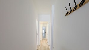 Egy keskeny folyosó, fa padlóval és fehér falakkal. A jobb oldali falon kabátakasztó található, a folyosó végén pedig egy ajtó.