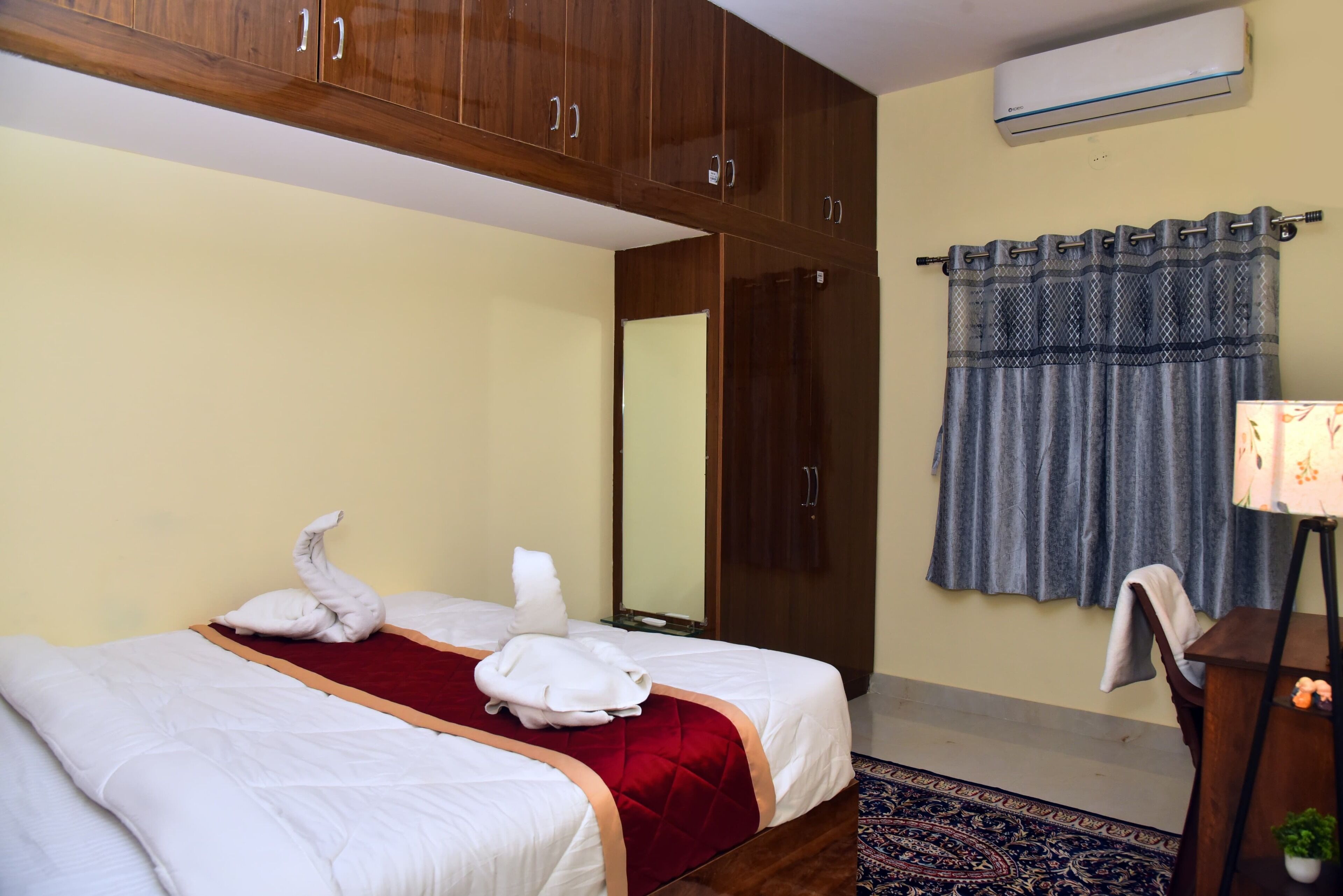 A hotel room with two beds, wooden wardrobes, a mirror, a window with curtains, and an air conditioning unit.