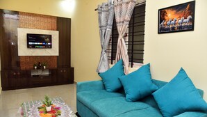 A living room with a teal sofa, a flat-screen TV mounted on a wooden panel, a patterned coffee table, and a painting of horses. - Tech Park Residency (Bengaluru)