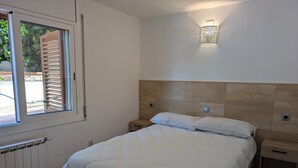 A bedroom with a bed, a nightstand, a wall-mounted light, a window with blinds, and a radiator.