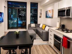 A modern kitchen with a dining area, a gray sofa with cushions, a black dining table, and a city view through large windows.