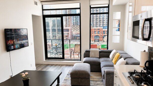A modern apartment with a large living area, a dining table, a kitchen with a stove, and large windows offering views of the city. - Downtown Calgary Haven - Steps to Stampede (Calgary)