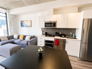 A modern kitchen with white cabinets, a stainless steel refrigerator, and a dining area with a black table. - Downtown Calgary Haven - Steps to Stampede (Calgary)