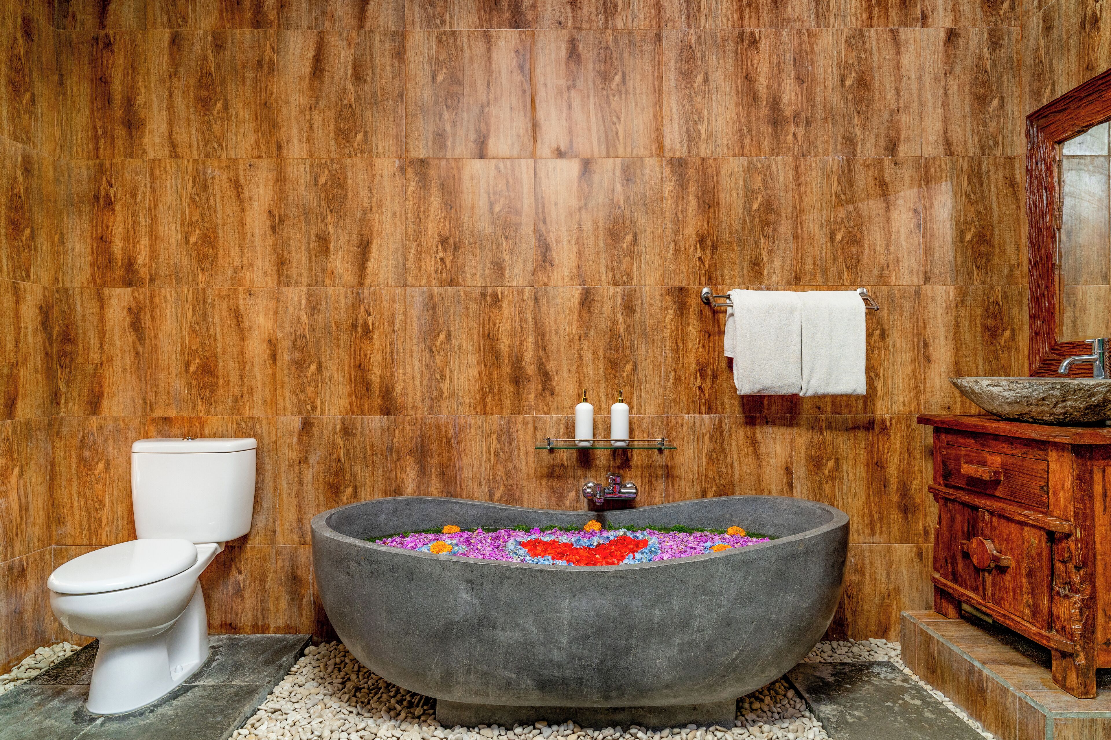 A bathroom with a wooden wall, a white toilet, a large bathtub filled with colorful flowers, and a wooden vanity with a basin.