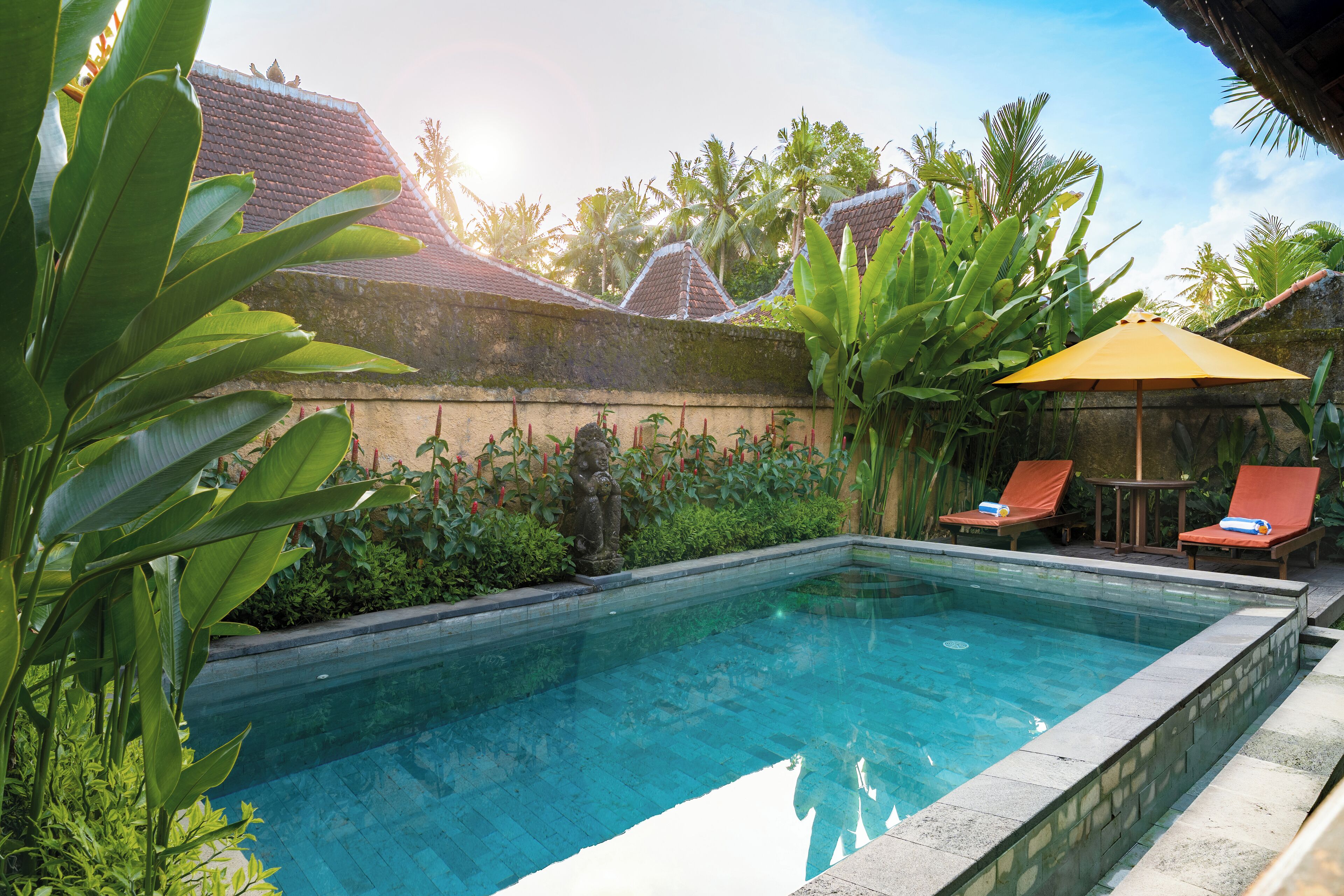 A rectangular pool with clear blue water, surrounded by lush greenery and a stone wall. There are two lounge chairs with orange cushions and a yellow umbrella.