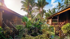 Un edificio tradizionale in legno con tetto di paglia, circondato da rigogliosa vegetazione tropicale e un piccolo stagno con una statua di Buddha.