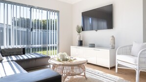 A modern living room with a sofa, a round coffee table, and a white armchair. A flat-screen TV is mounted on the wall, and there is a large window with blinds.