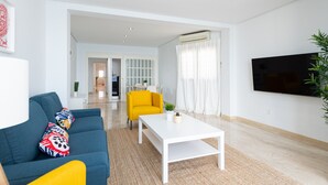 Una sala de estar moderna con un sofá azul, una mesa de centro blanca y un sillón amarillo. Hay un televisor de pantalla plana montado en la pared y una planta en maceta a la derecha.
