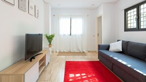 Una sala de estar moderna con un televisor de pantalla plana, un sofá gris, una alfombra roja y un ventanal con cortinas blancas.