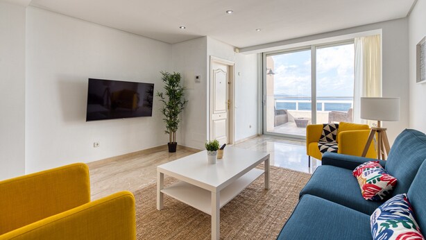 Una sala de estar moderna con un televisor de pantalla plana, una mesa de centro blanca, un sofá azul, sillones amarillos y un ventanal con vistas.