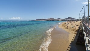 A beach with clear water, a sandy shore, and a cityscape in the background.