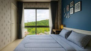 Une chambre avec un grand lit, une table de chevet, une lampe et une vue sur la verdure et les bâtiments, visible par une fenêtre.