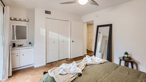 Une chambre à coucher comprenant un lit, une coiffeuse avec un miroir, un ventilateur de plafond et une porte donnant sur une autre pièce.
