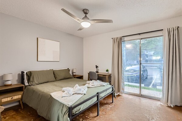 A bedroom with a bed, a desk, a chair, a ceiling fan, and a window with curtains.